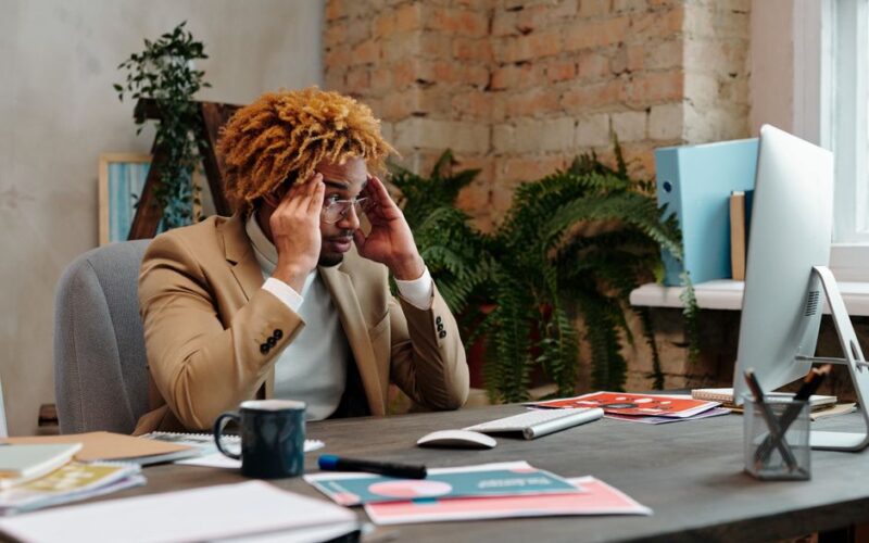 A businessman in stress at his office desk, overwhelmed with work documents and computer monitor.