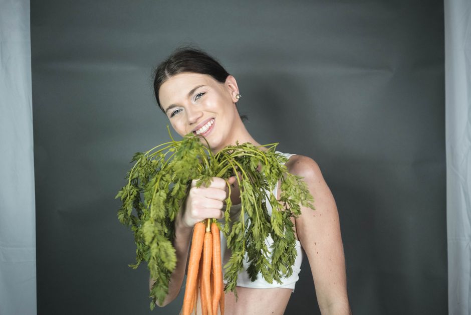 A cheerful woman poses with a bunch of fresh carrots against a gray background in a studio setting.