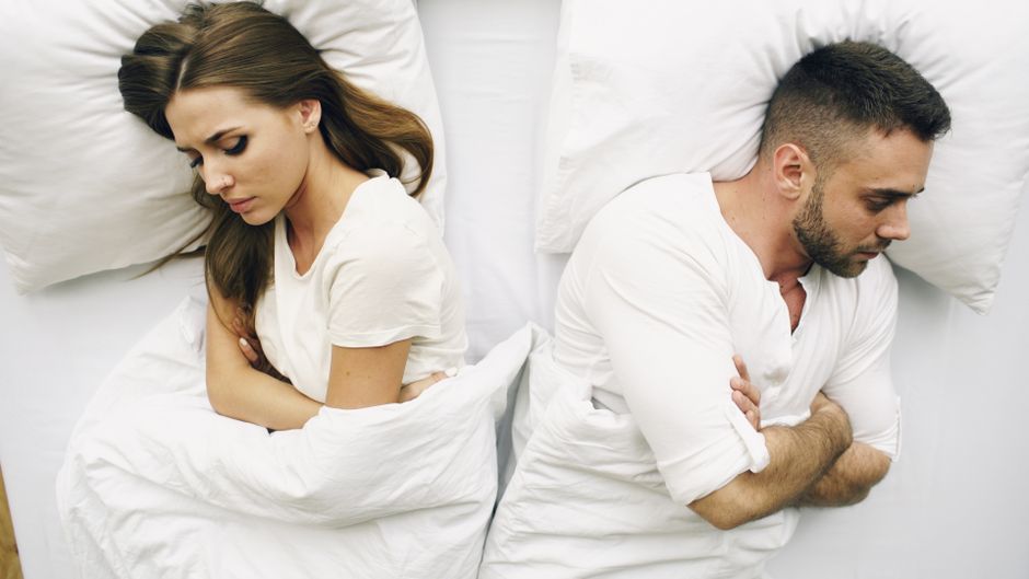 A couple lying in bed facing away, expressions of discontent.