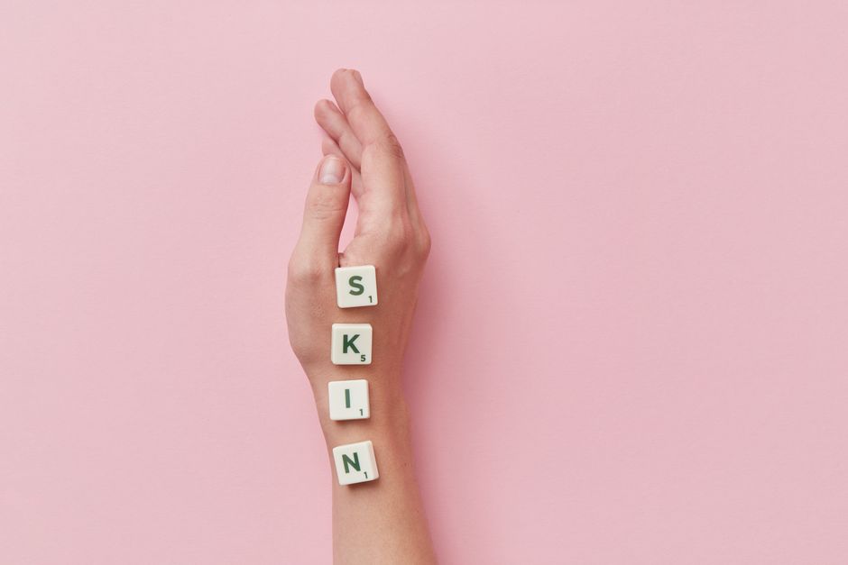 A creative arrangement of Scrabble tiles spelling 'skin' on a hand against a pink backdrop.