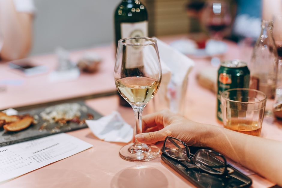A hand holding a wine glass at a casual dining table with various beverages and a smartphone.