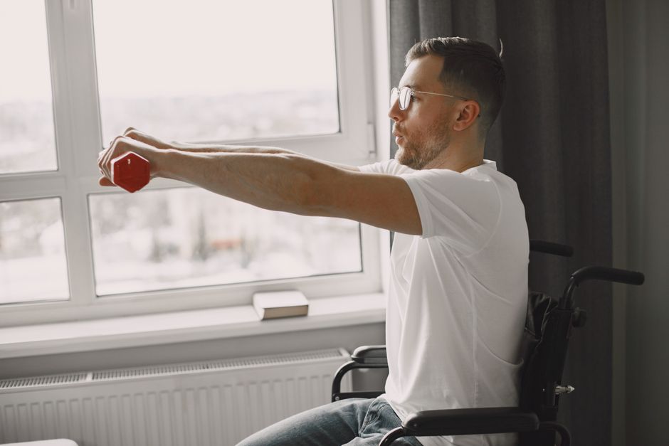 A man in a wheelchair exercising with a dumbbell by a bright window, promoting inclusivity and health.