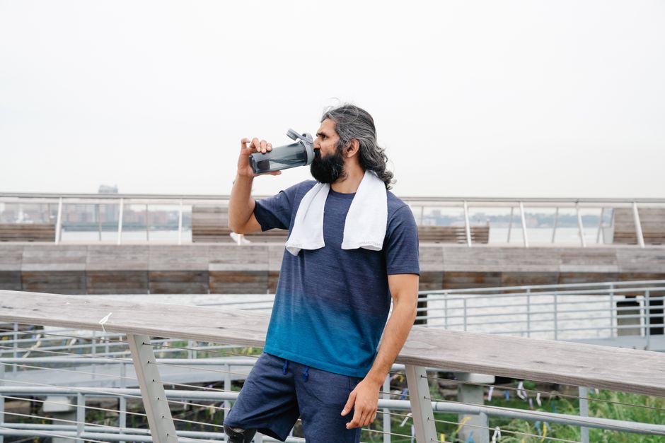 A man takes a refreshing break, drinking water outdoors after a workout, with a towel draped over his shoulders.