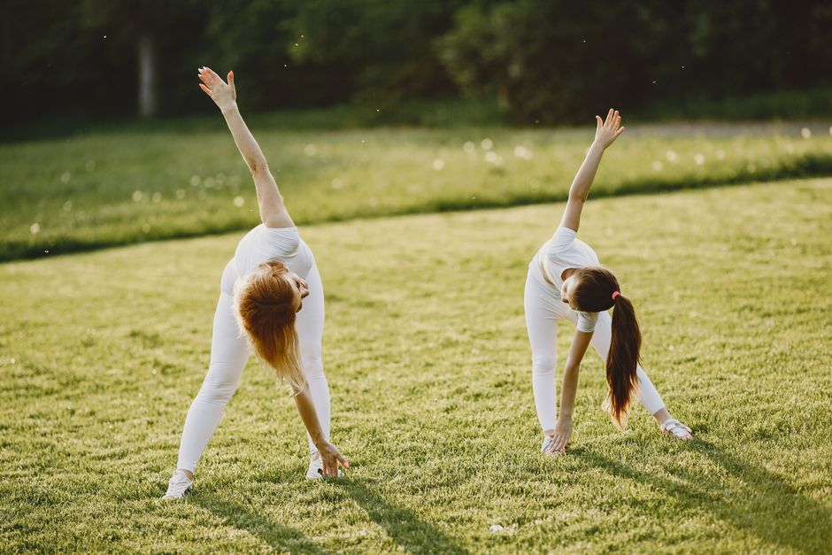A mother and daughter bond while stretching in a sunny grass field, dressed in white activewear.