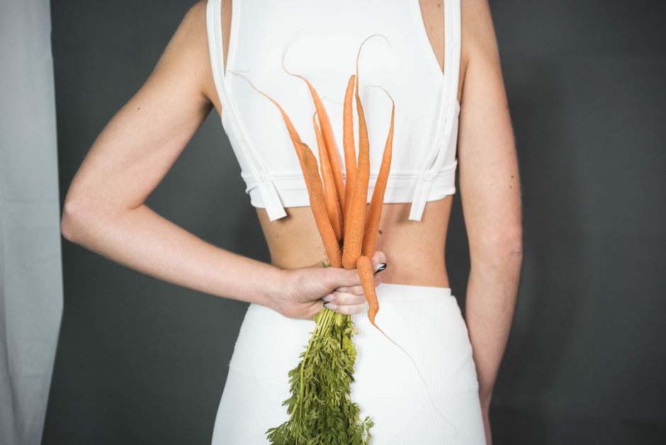 A person in white holding a bunch of fresh carrots against a gray background, symbolizing health and wellness.