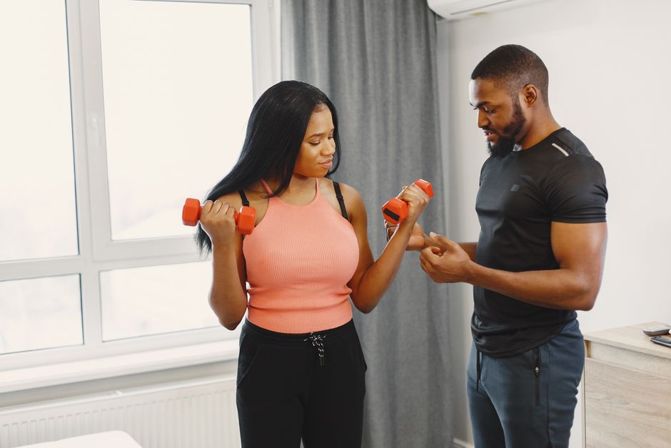 A personal trainer guides a woman during her dumbbell workout in a bright home gym.
