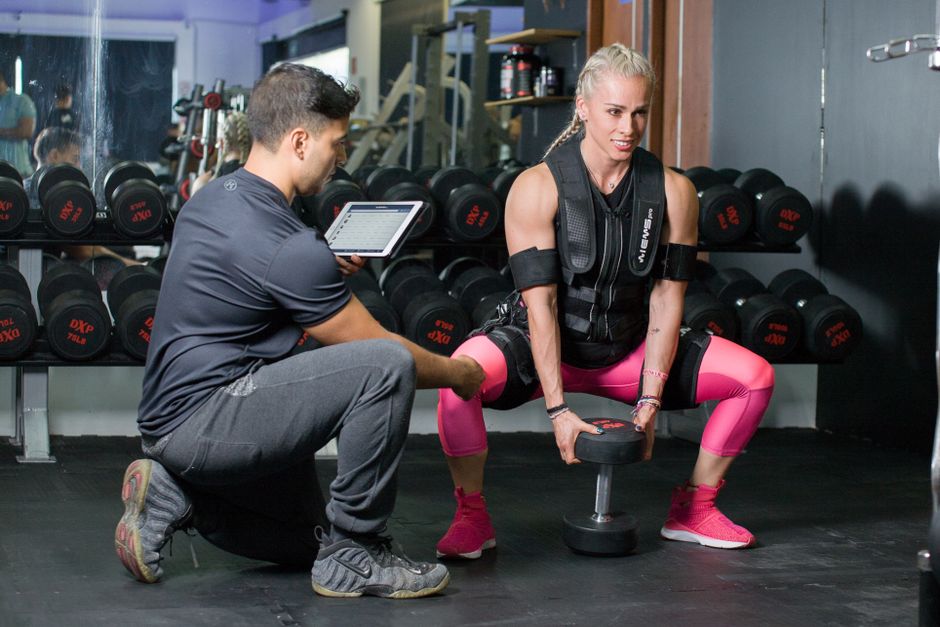A personal trainer guides a woman performing a dumbbell squat in a gym setting.