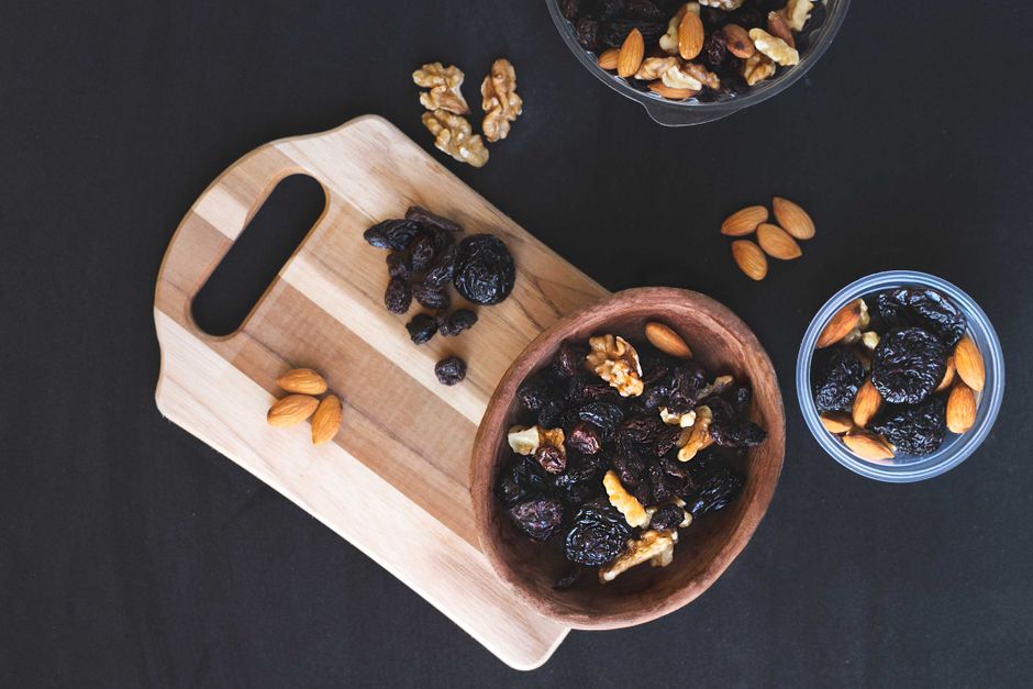 A top-down view of mixed dried fruits and nuts in bowls on a black surface with a wooden chopping board.