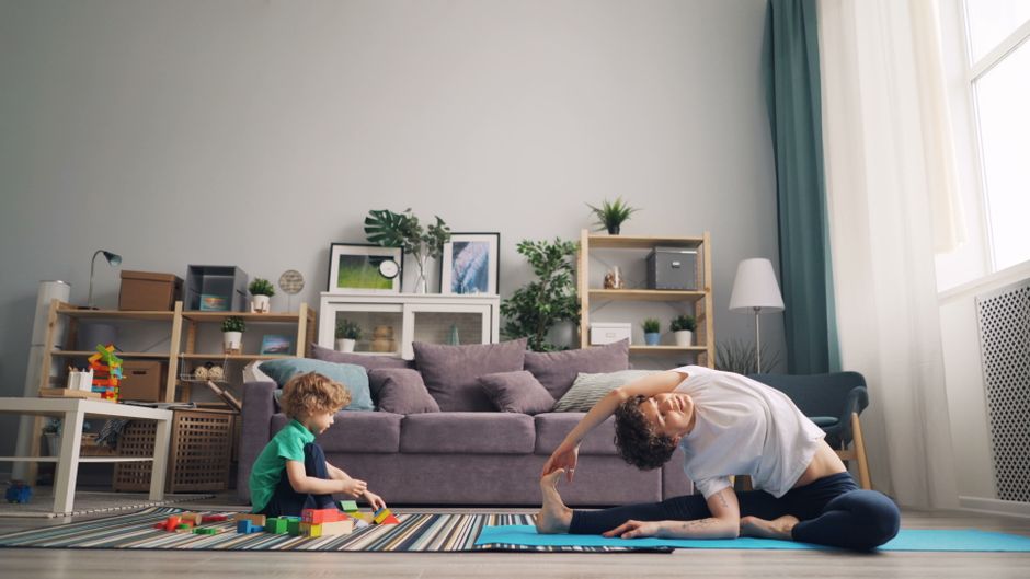 A woman practicing yoga at home while her son plays with blocks beside her on a mat.