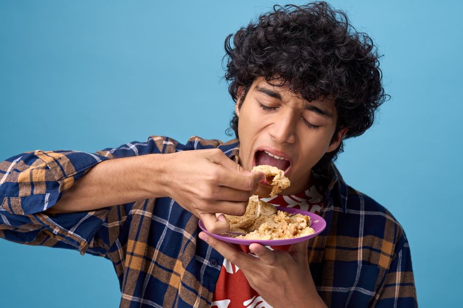 A young man eagerly eating Indian bread and curry against a blue background.