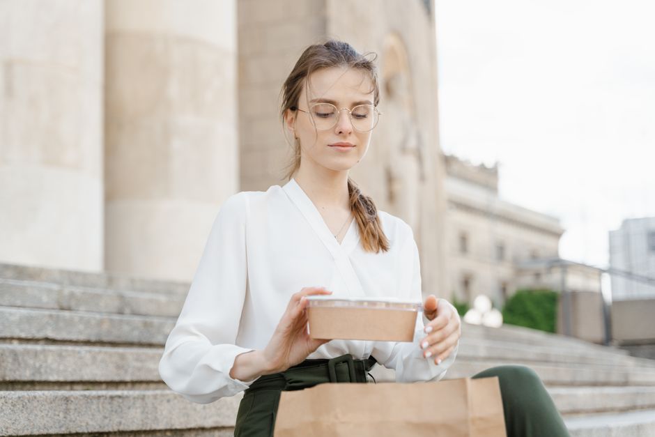 A young woman in a white blouse enjoys a takeaway lunch while sitting on urban steps.