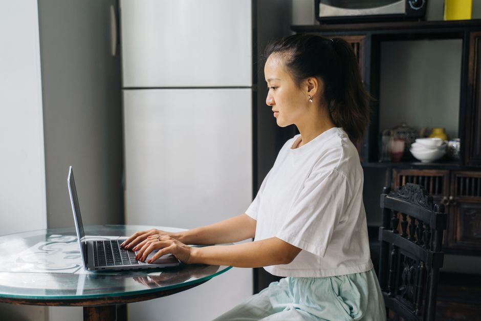 A young woman sits at a table working on a laptop in a home office setting.