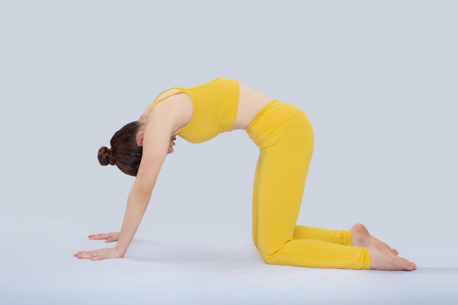 An adult woman in a yellow outfit performing cat pose yoga indoors, focusing on flexibility and well-being.