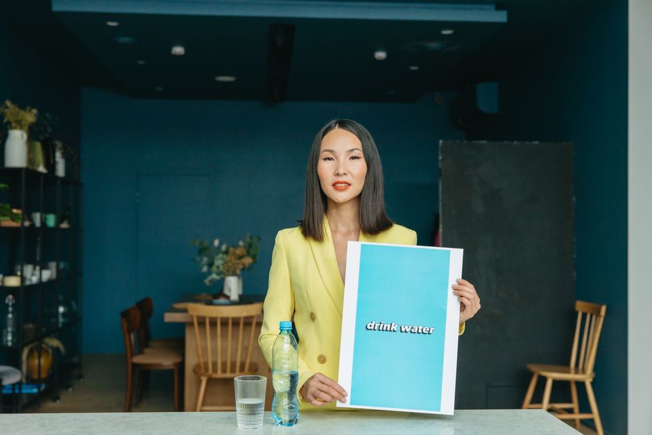 An Asian woman in a yellow blazer holds a "drink water" poster, emphasizing hydration in an office setting.