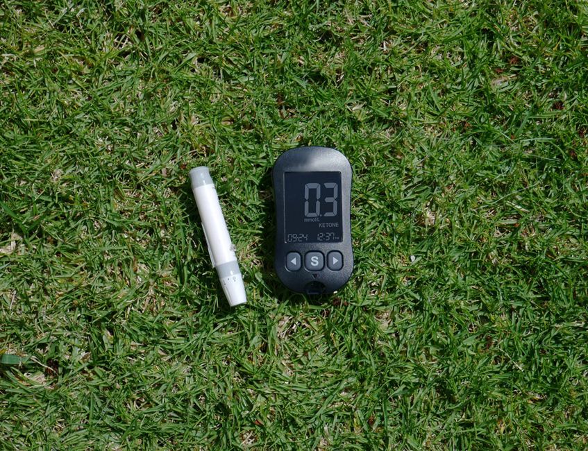 Blood ketone meter and strip displayed outdoors on grass.
