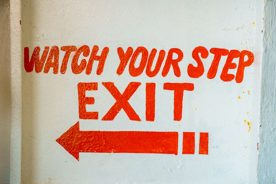 Bold red 'Watch Your Step' and 'Exit' sign with an arrow on a white textured wall.