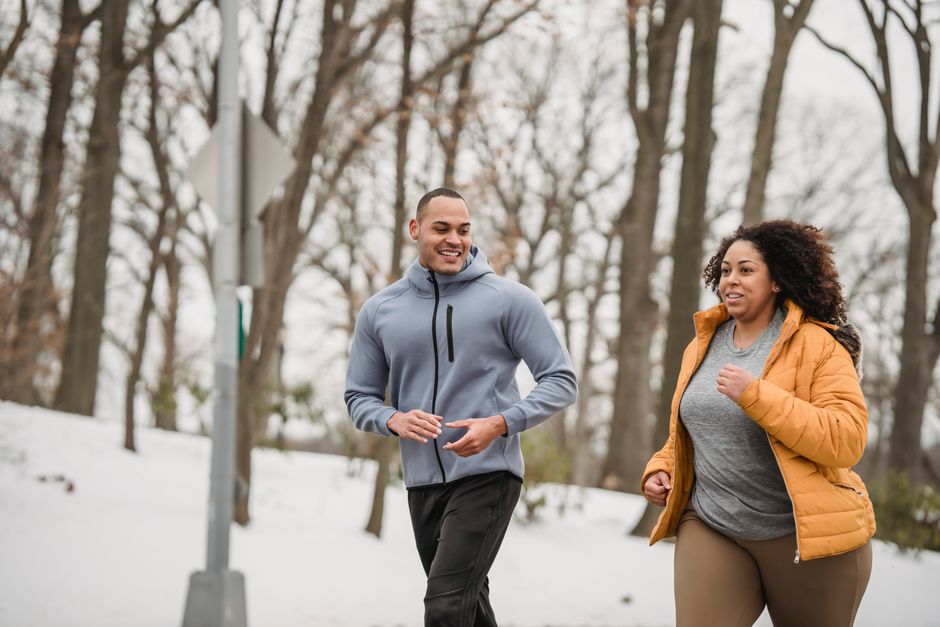 Cheerful sporty ethnic male trainer jogging with plus sized black female in warm jacket in snowy winter park