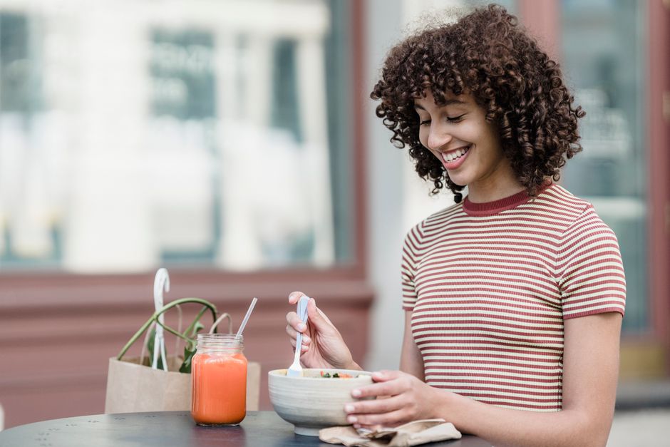 Cheerful young female with bowl of yummy food and jar of refreshing beverage sitting at table in urban cafeteria