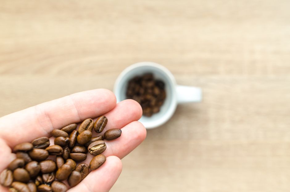 Close-up of a hand holding roasted coffee beans with a coffee cup in the background on a wooden table.