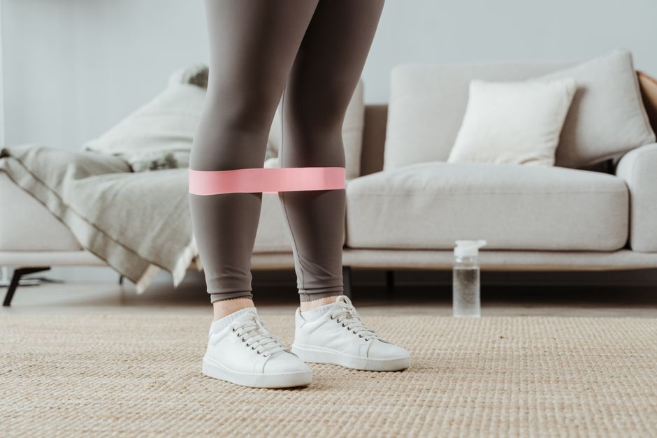 Close-up of a woman in sport leggings using a pink resistance band for leg exercises indoors.