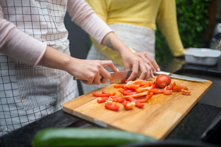 Close-up of hands slicing red bell peppers and tomatoes on a wooden cutting board in a kitchen setting.