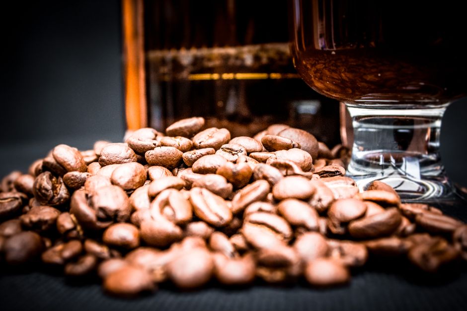 Closeup of pile of aromatic coffee beans with glass of coffee and blurred French press behind on black background
