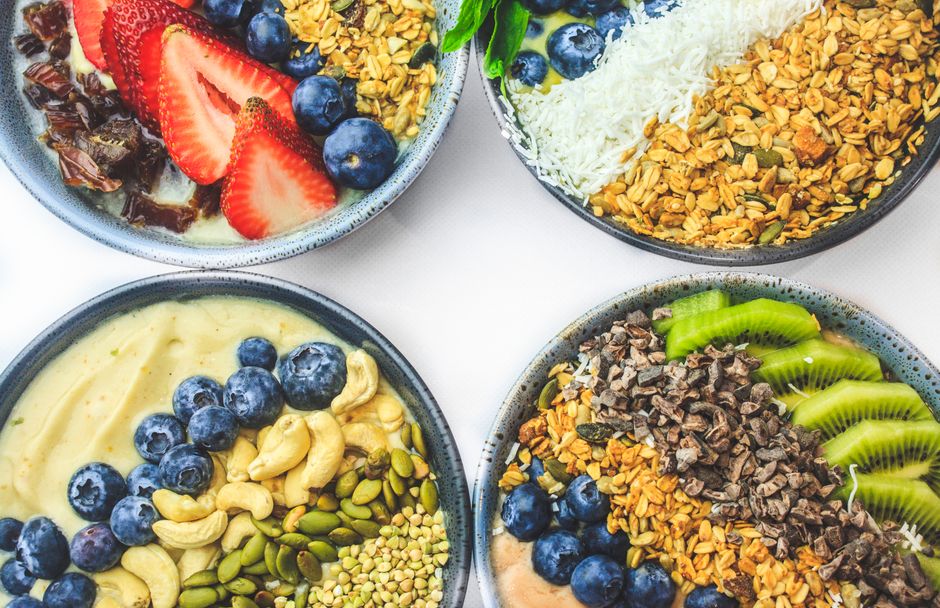 Colorful breakfast bowls with fresh fruits, granola, and seeds on a white surface.
