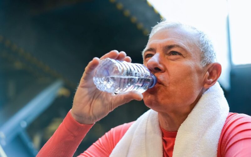 Elderly man drinking water post-exercise with towel around neck, promoting healthy lifestyle.