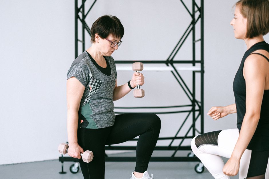 Elderly woman lifting dumbbells with guidance from a trainer indoors.