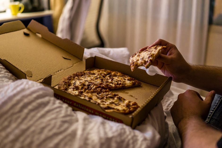 From above side view of crop anonymous male eating tasty slice of pizza with cheese in takeaway cardboard box on crumpled blanket in bed in apartment