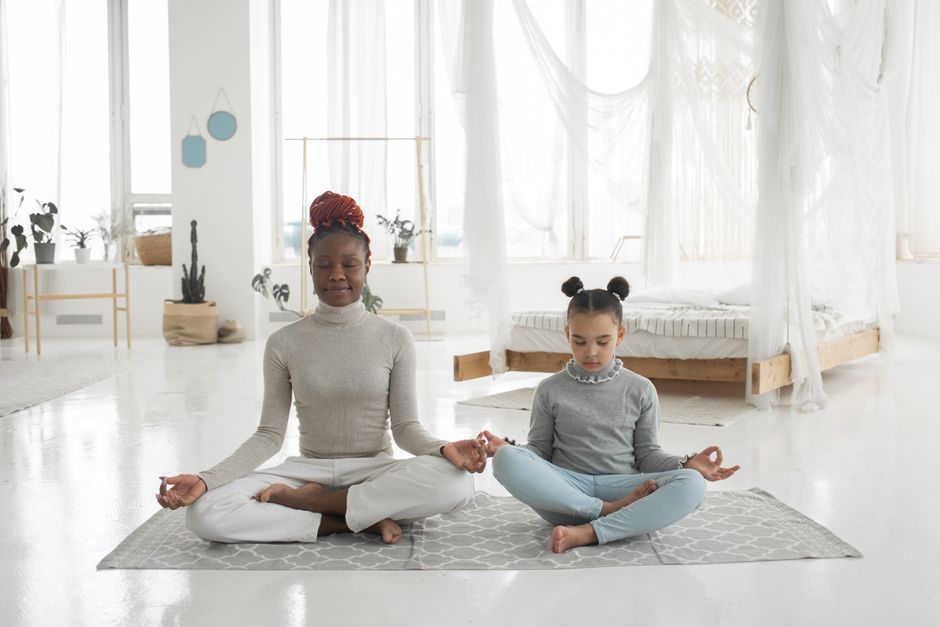 Full body of concentrated young barefooted black woman with cute little daughter sitting together on floor in Padmasana pose and mudra hands and meditating with closed eyes in light bedroom