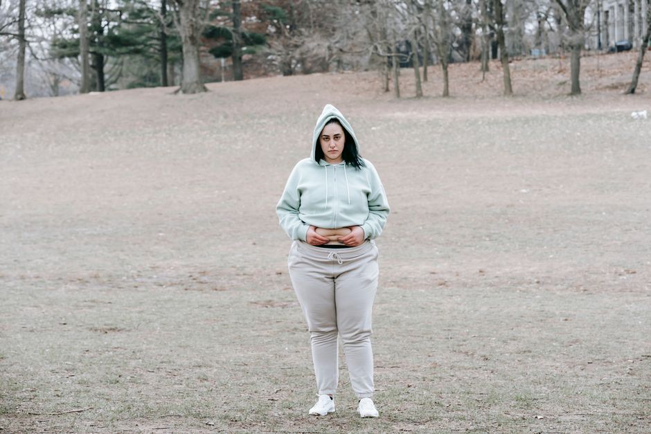 Full length of unhappy young woman in activewear standing in autumn park and showing fat on waist