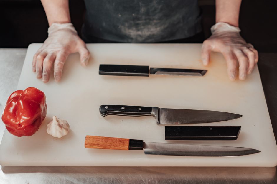 Gloved hands preparing to chop with knives on a cutting board next to vegetables in a kitchen scene.