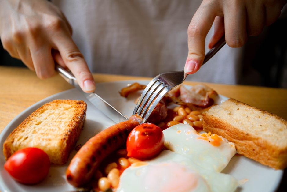 Hands preparing a hearty full English breakfast with eggs, beans, sausage, and toast.