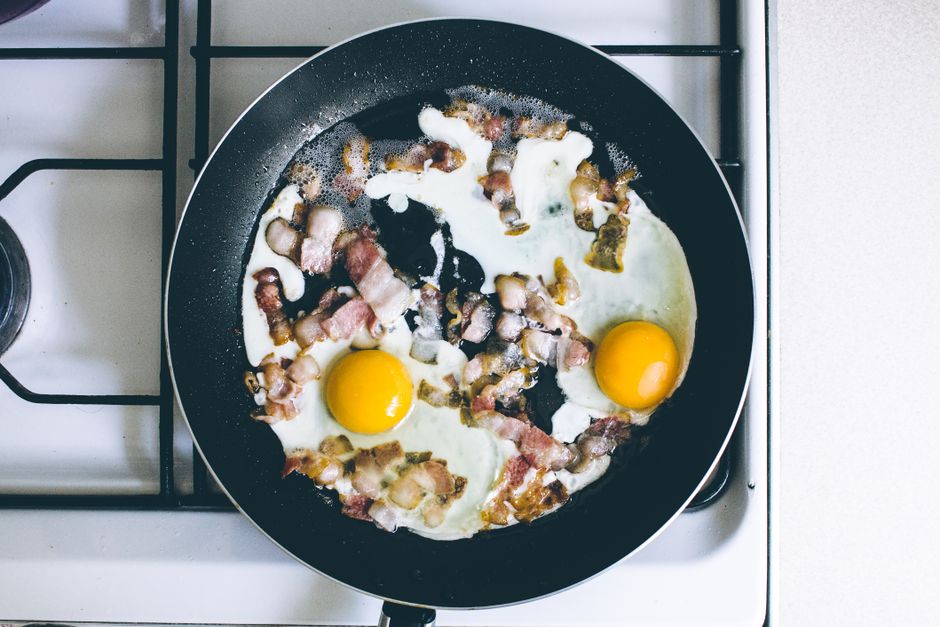 High-angle view of fried eggs and bacon sizzling in a skillet on a gas stove, perfect breakfast scene.