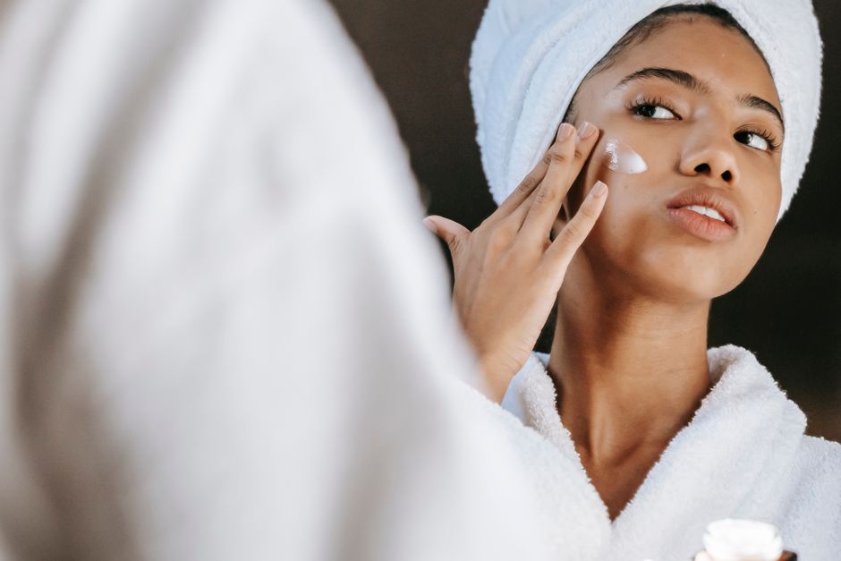 Low angle of ethnic female in towel looking at mirror while treating skin with moisturizing lotion during morning routine
