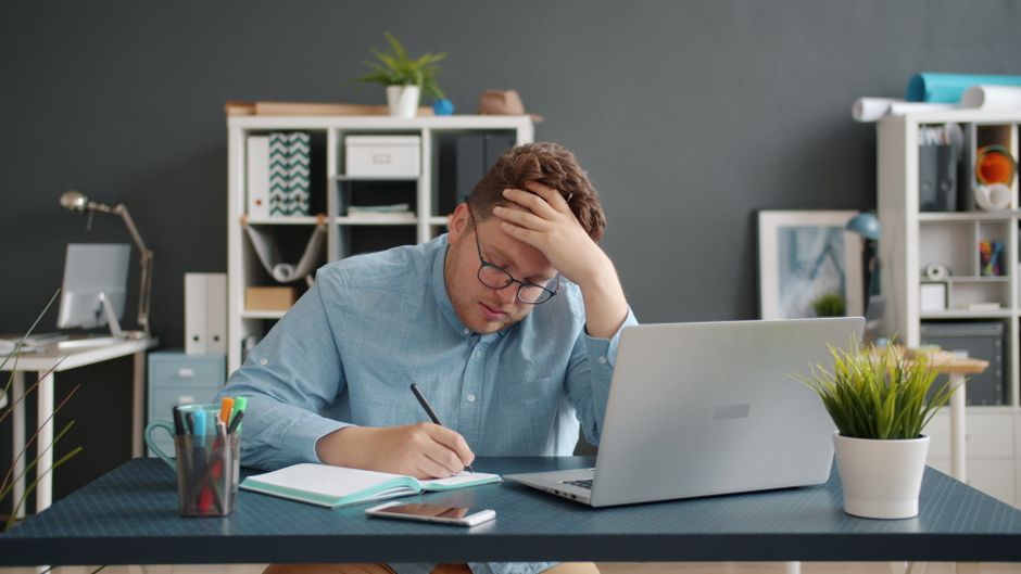 Man in office writes notes by laptop, looks stressed. Modern workspace.