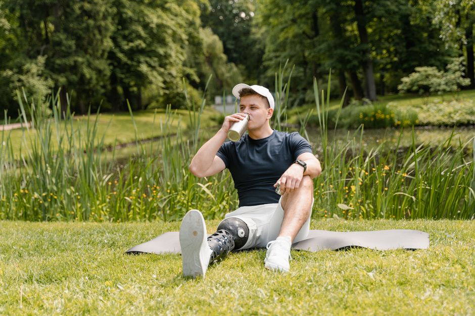 Man with a prosthetic leg drinks water on a mat in a tranquil park setting.
