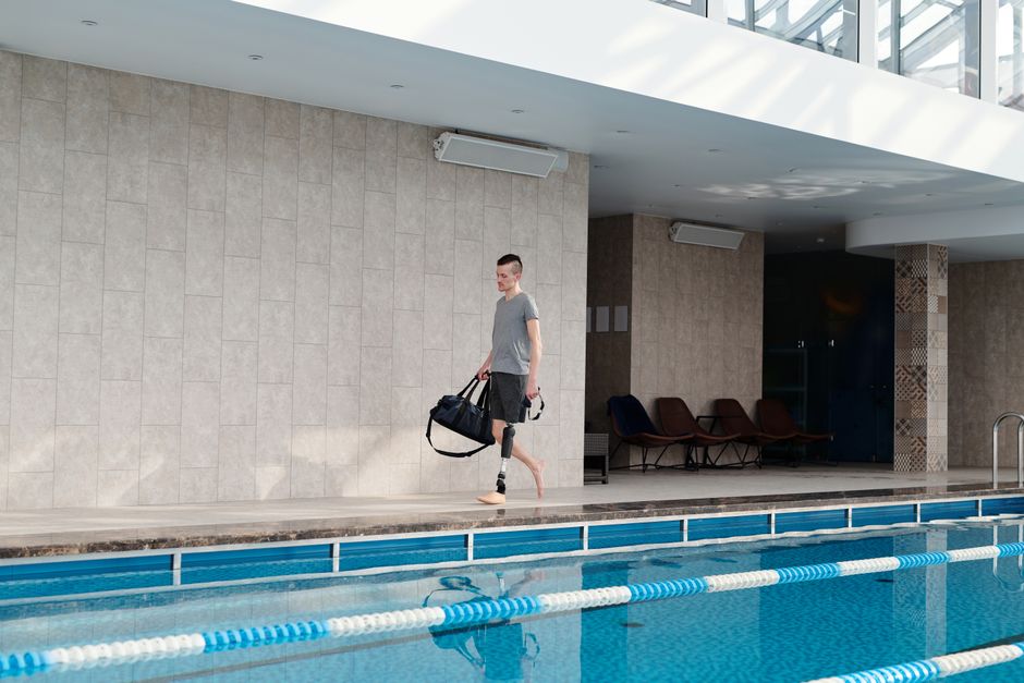 Man with prosthetic leg walking beside an indoor swimming pool carrying a gym bag.