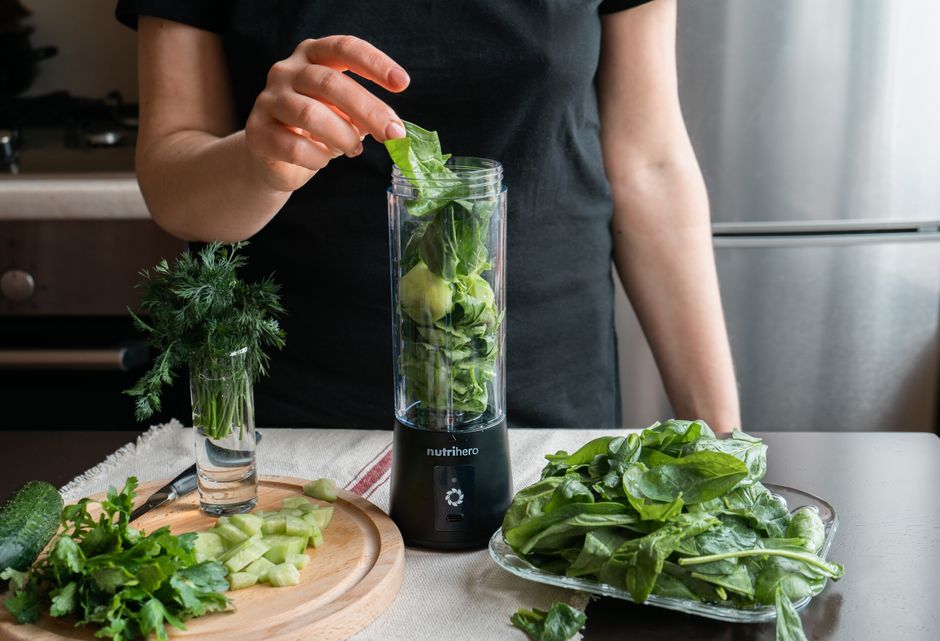 Person preparing a fresh green smoothie with spinach, cucumber, and herbs using a blender.