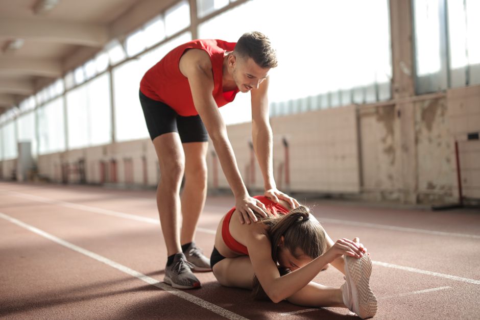 Professional male sportsman helping woman touching knees with head while female sitting on running track and doing stretching exercise