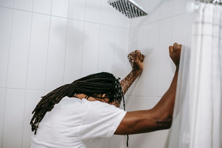 Side view of anonymous ethnic male with dreadlocks and mental problem touching ceramic wall under water fluid in shower cabin