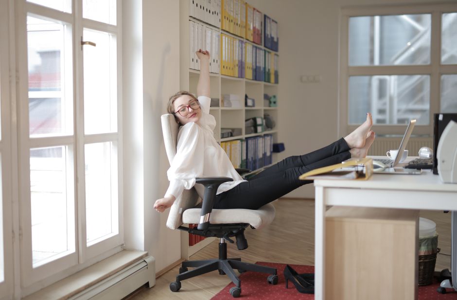 Side view of satisfied young female assistant wearing classy clothes and eyeglasses sitting barefoot on office chair with feet on table and stretching with closed eyes
