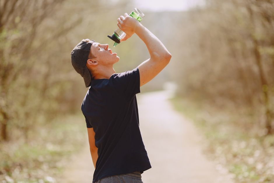 Side view of young male athlete wearing sports clothes and cap drinking water from plastic bottle on blurred background of forest during running and workout