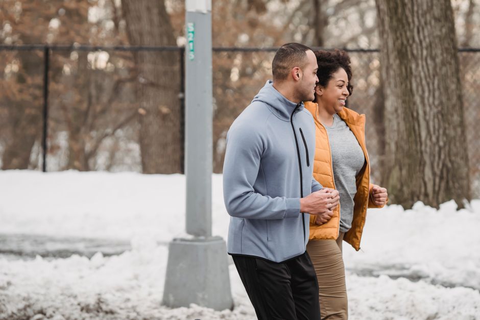 Side view smiling ethnic male trainer and plus sized black female in warm clothes running together in snowy winter park
