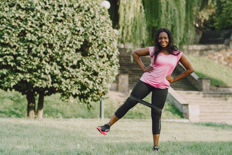 Smiling woman doing leg workouts with resistance band in a sunny park.