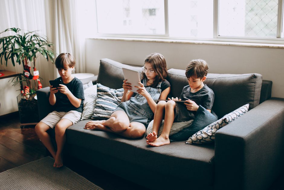 Three children on a gray sofa engrossed in digital devices, indoors.