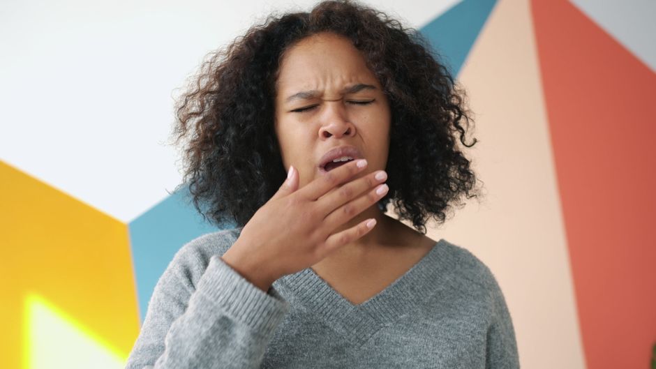 Tired young woman yawning, standing against colorful geometric wall indoors.