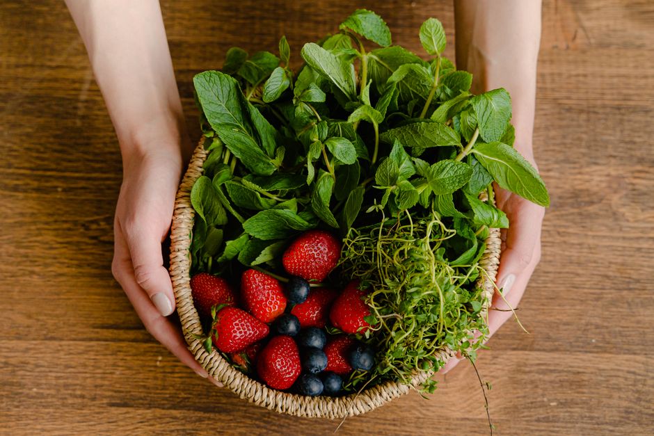 Top view of a basket filled with fresh strawberries, blueberries, and mint, held by hands on a wooden table.