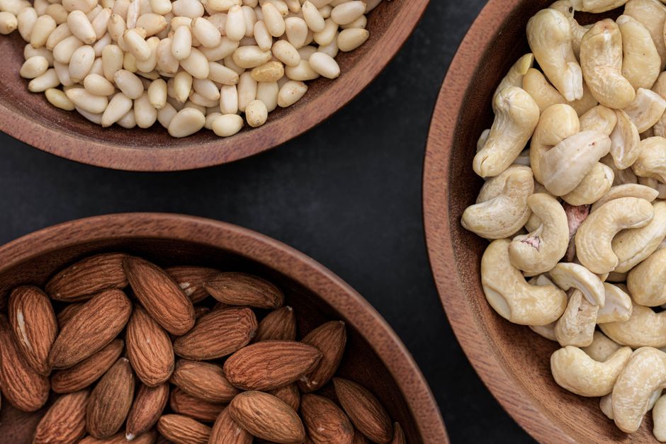Top view of almonds, cashews, and pine nuts in wooden bowls against a dark backdrop.